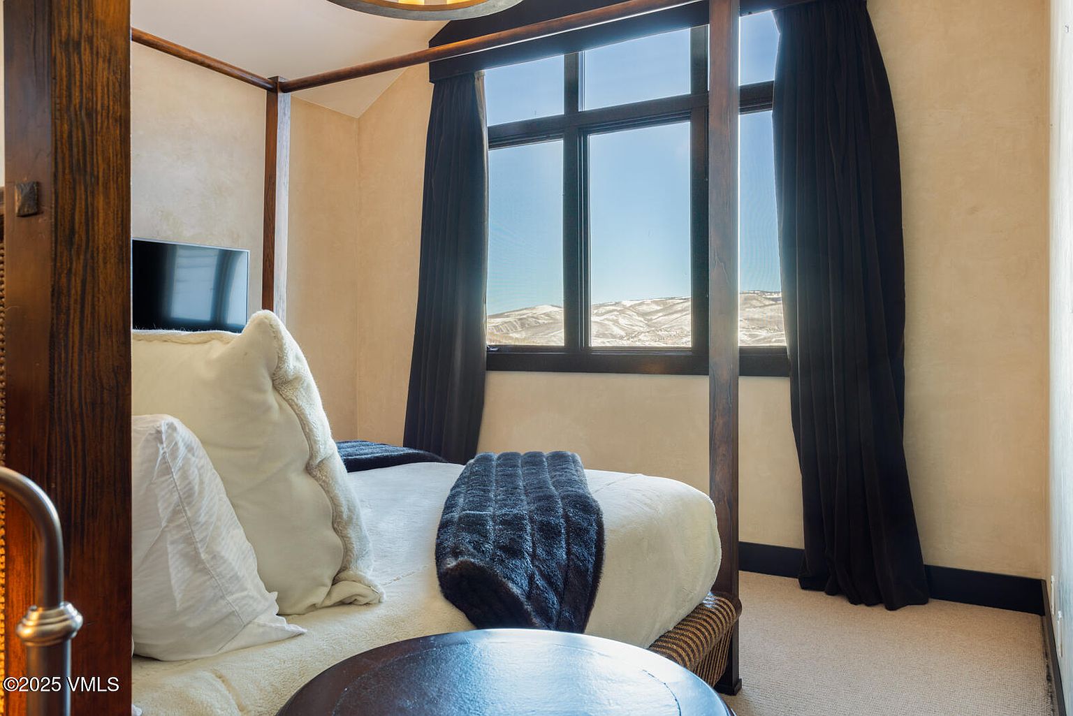 This is a cozy bedroom featuring a four-poster bed with dark wood framing. The bed is adorned with white linens, pillows, and a dark blue throw blanket. A window with dark curtains provides natural light and a view of snow-covered mountains in the distance, creating a serene and inviting atmosphere.