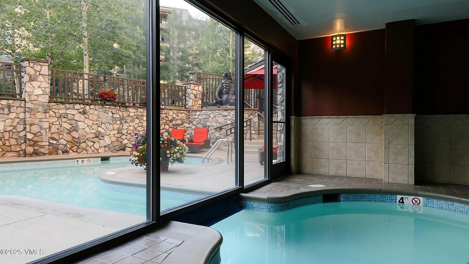 This image showcases a luxurious indoor-outdoor pool and spa area. The indoor spa features tiled walls and a clear view through large windows to an outdoor pool area with stone walls, landscaping, and lounge chairs. The scene conveys a sense of relaxation and upscale living.