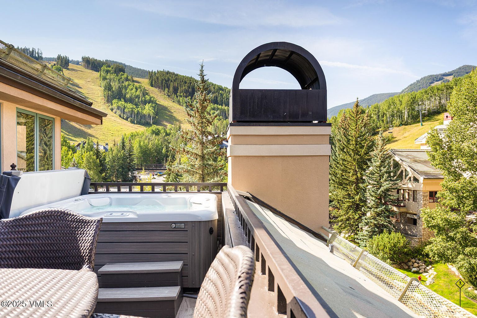This image showcases a luxurious patio/deck/balcony featuring a hot tub with steps for easy access. The space is furnished with wicker chairs and a table, creating a relaxing outdoor living area. The backdrop includes scenic mountain views and lush greenery, enhancing the property's appeal.