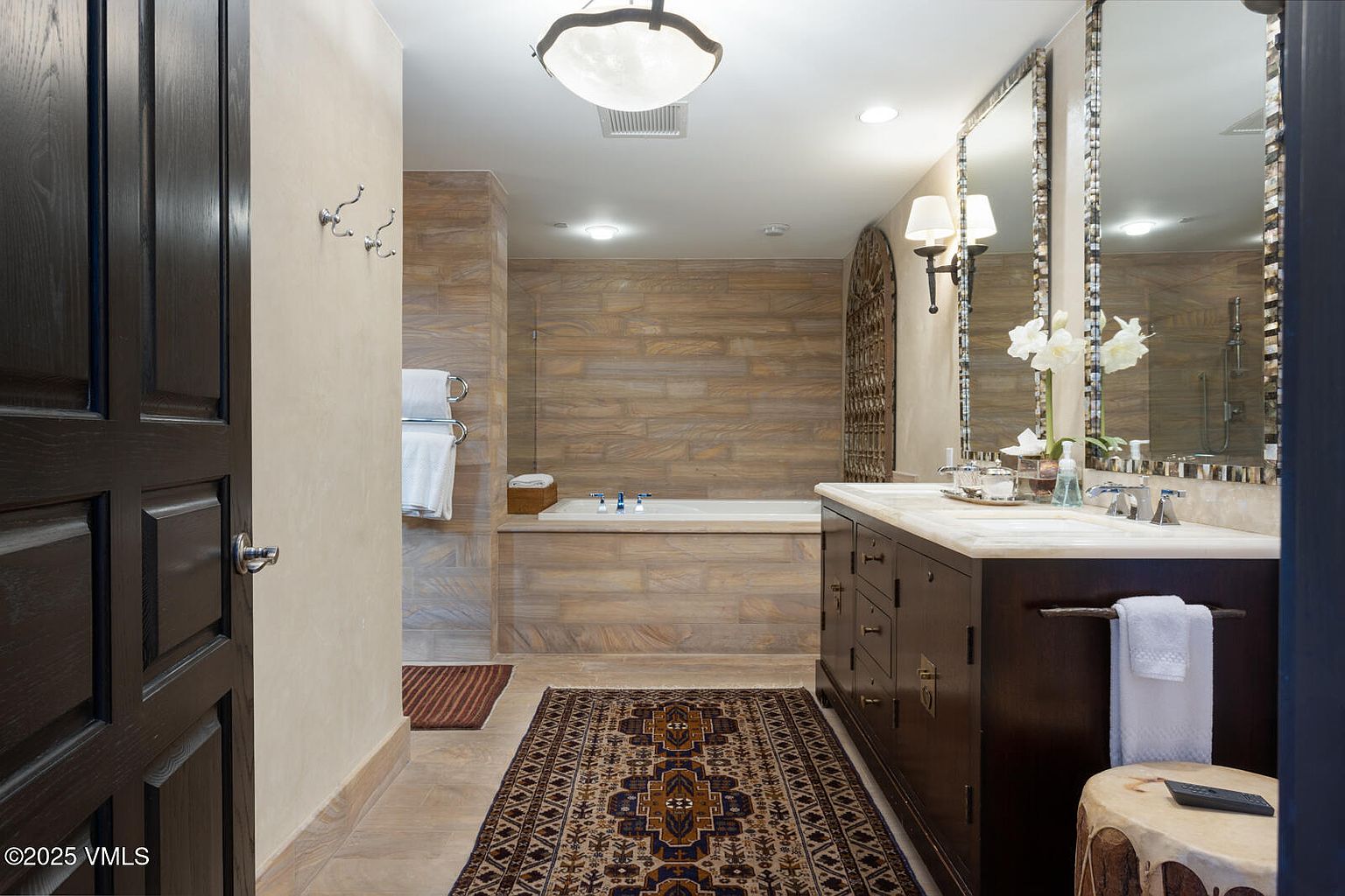 This is a primary bathroom featuring a dark wood vanity with a marble countertop, dual sinks, and decorative mirrors. A bathtub is visible in the background, surrounded by horizontally laid stone tiles that extend to the walls. An ornate rug adds warmth to the tiled floor, and the overall design exudes a luxurious and spa-like atmosphere.