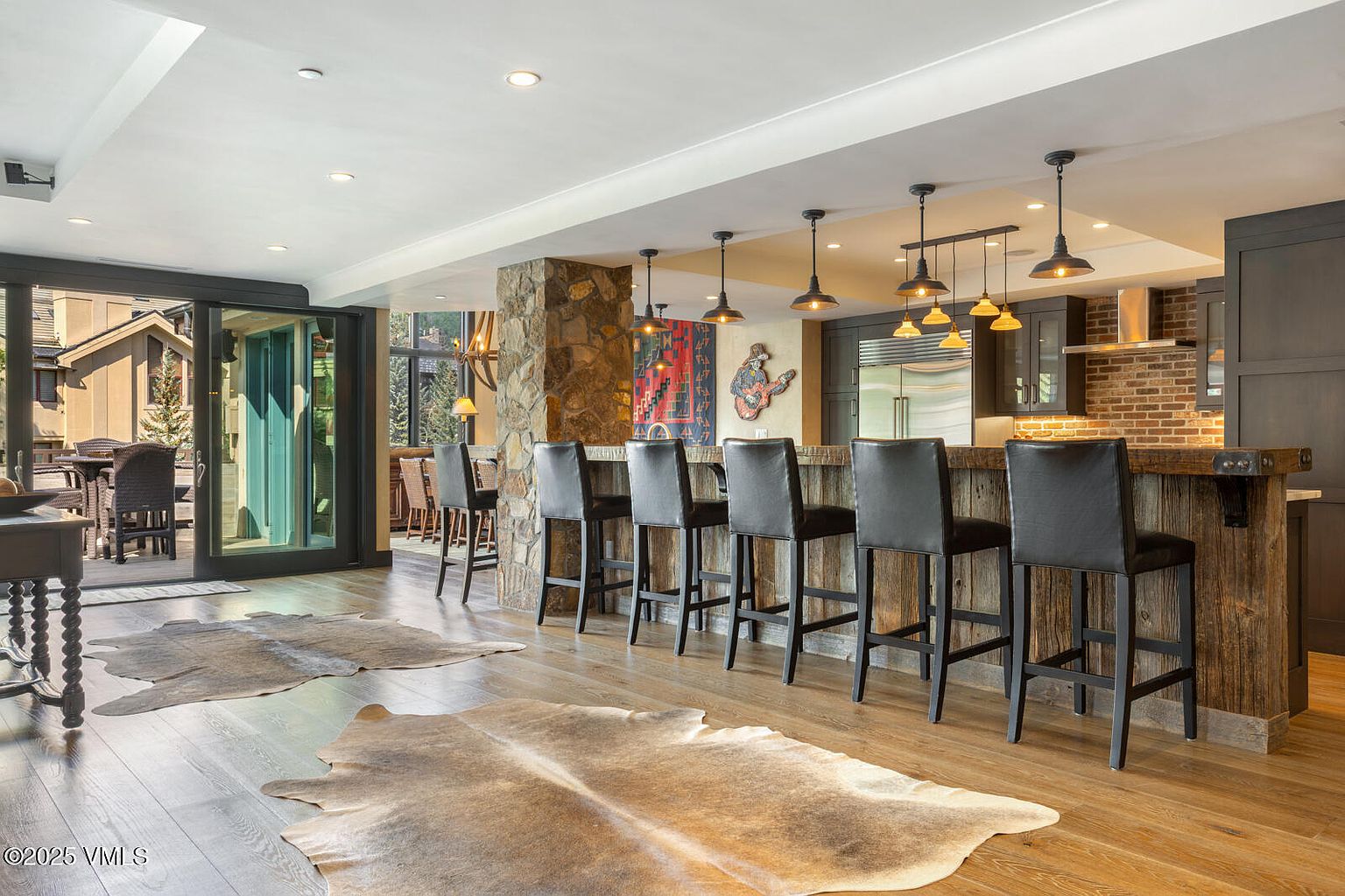 This interior shot showcases a stylish kitchen area with a rustic-modern design. The kitchen features a wooden bar with black leather bar stools, complemented by pendant lighting and a stone pillar. Cowhide rugs add a touch of warmth to the hardwood floors, while a sliding glass door leads to an outdoor patio area.