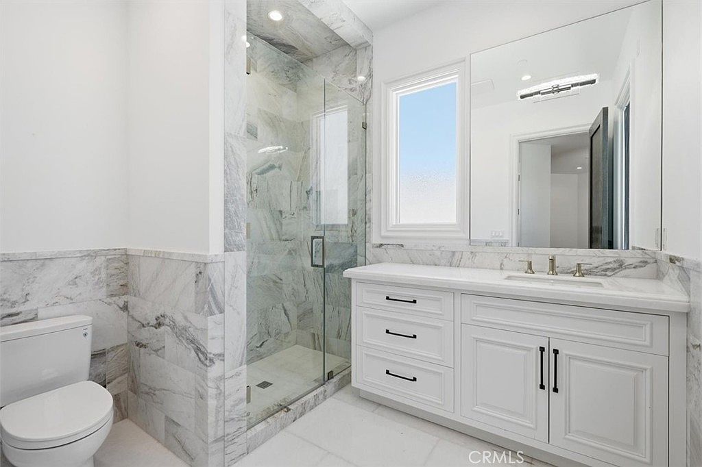 This bright, modern bathroom features a white vanity with a stone countertop and a glass-enclosed shower with patterned stone walls. A large mirror sits above the vanity, reflecting the space and a window that allows for natural light. The room is finished with light-toned tile flooring and a white toilet, creating a clean and cohesive aesthetic.