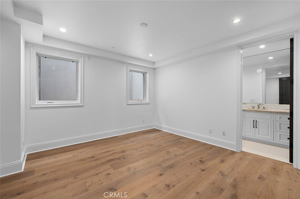 This bright, minimalist bedroom features light-toned wood flooring and crisp white walls, creating a clean and airy atmosphere. Two windows on the left wall provide natural light, while an open doorway on the right leads into an adjoining bathroom with a vanity and mirror. The room is unfurnished, highlighting the neutral palette and modern recessed lighting throughout the ceiling.