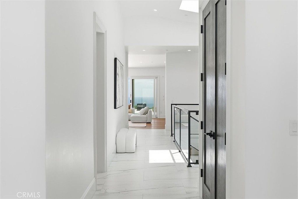 This bright, minimalist hallway features white walls and light-toned stone flooring, creating an airy and expansive atmosphere. A dark-framed door stands to the right, while a glass-paneled railing leads toward a staircase, offering a view into a sunlit living area with a distant ocean vista. The perspective draws the eye through the clean, modern corridor toward the open space beyond.