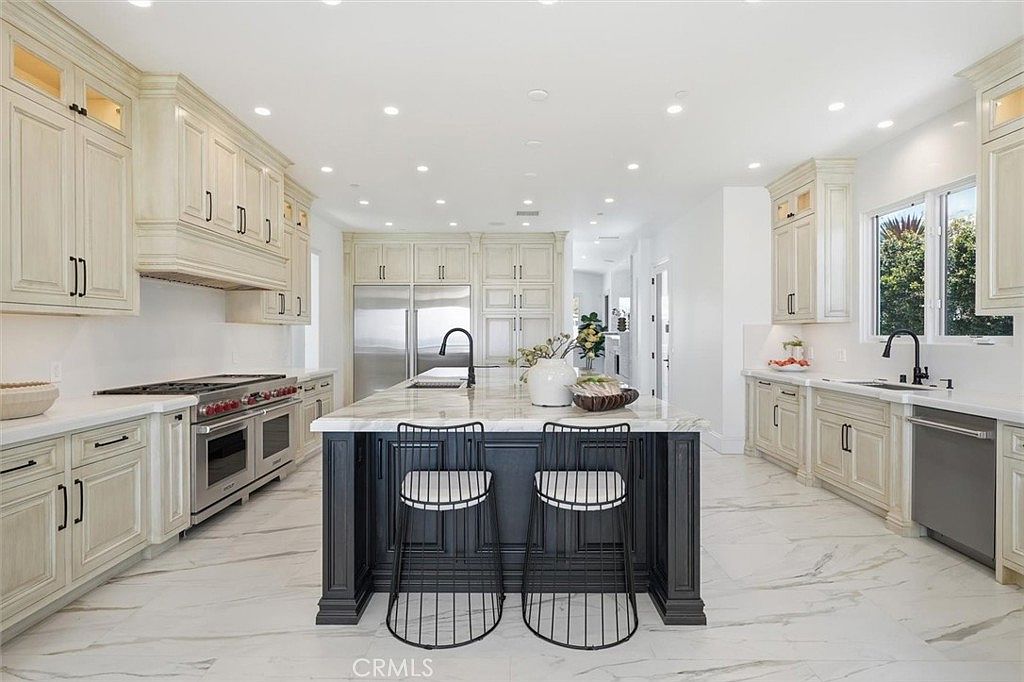 This bright and spacious kitchen features a large central island with a dark base and stone countertop, complemented by two wire-frame bar stools. The perimeter is lined with light-toned cabinetry, a professional-grade range, and a secondary sink area with a dishwasher. The room is finished with light-colored stone flooring and recessed lighting, creating a clean and sophisticated aesthetic.