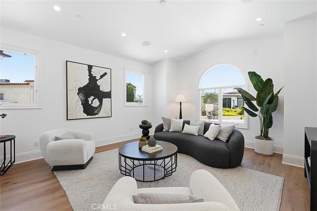 This bright and airy living room features a contemporary design with a curved charcoal sofa, two plush white armchairs, and a round coffee table centered on a textured area rug. The space is accented by a large abstract piece of wall art and a tall potted plant, while light wood flooring runs throughout. Large windows, including an arched window, allow for ample natural light and provide a view of the outdoor landscape.