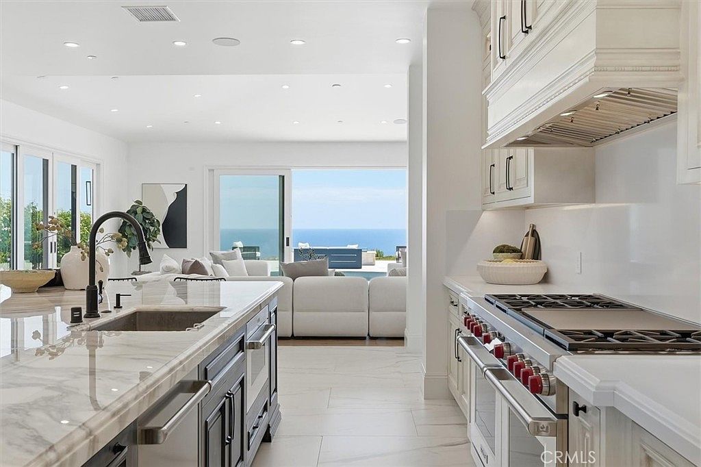 This bright, modern kitchen features a large central island with a stone countertop and a dark-toned base, complemented by crisp white cabinetry and a professional-grade range. The open-concept design flows seamlessly into a living area, where large sliding glass doors reveal a stunning, unobstructed view of the ocean. The space is characterized by its clean lines, neutral color palette, and an airy, sophisticated atmosphere that emphasizes indoor-outdoor living.