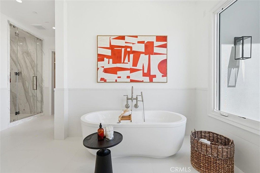 This bright and modern primary bathroom features a freestanding soaking tub as the central focal point, positioned beneath a large piece of abstract orange and white wall art. To the left, a glass-enclosed walk-in shower with light-toned stone walls adds a luxurious touch, while a woven basket and a small side table with toiletries complete the serene aesthetic. The space is illuminated by natural light from a large window on the right, creating an airy and spa-like atmosphere.