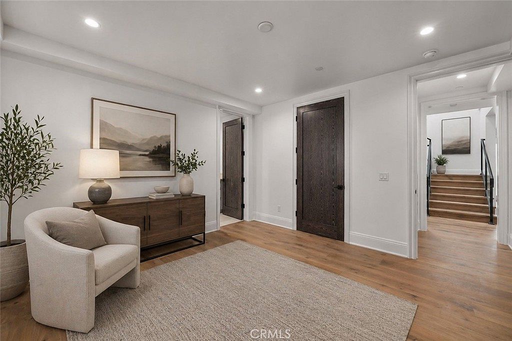 This bright and airy hallway features light wood flooring and crisp white walls, creating a clean, modern aesthetic. A console table with a lamp and artwork sits against the wall, while a comfortable armchair provides a cozy seating area on the textured rug. The space leads toward a staircase in the background, with dark-toned doors adding a sophisticated contrast to the neutral palette.
