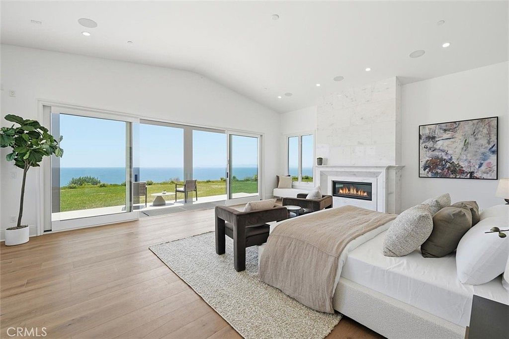 This spacious primary bedroom features light wood flooring and a neutral color palette, centered around a large bed with plush bedding. A tall stone-clad chimney breast houses a modern fireplace, while expansive glass sliding doors provide a seamless transition to the outdoor patio and offer panoramic ocean views. The room is bright and airy, complemented by recessed lighting and a piece of abstract wall art.