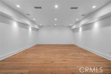 This spacious, minimalist room features light-toned wood flooring and crisp white walls, creating a bright and airy atmosphere. The ceiling is equipped with recessed lighting and integrated speakers, suggesting a versatile space suitable for a media room or recreational area. The perspective is centered, looking across the expansive, open floor plan toward the back wall.