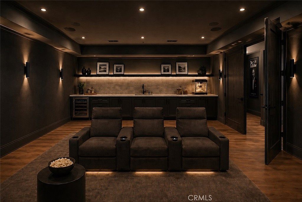 This sophisticated home theater features three plush, dark-toned reclining seats arranged on a neutral area rug. Behind the seating area, a built-in wet bar with dark cabinetry, a sink, and a popcorn machine is illuminated by warm under-cabinet lighting. The room is finished in a moody, dark color palette with recessed ceiling lights and wall-mounted sconces, creating an intimate and cinematic atmosphere.