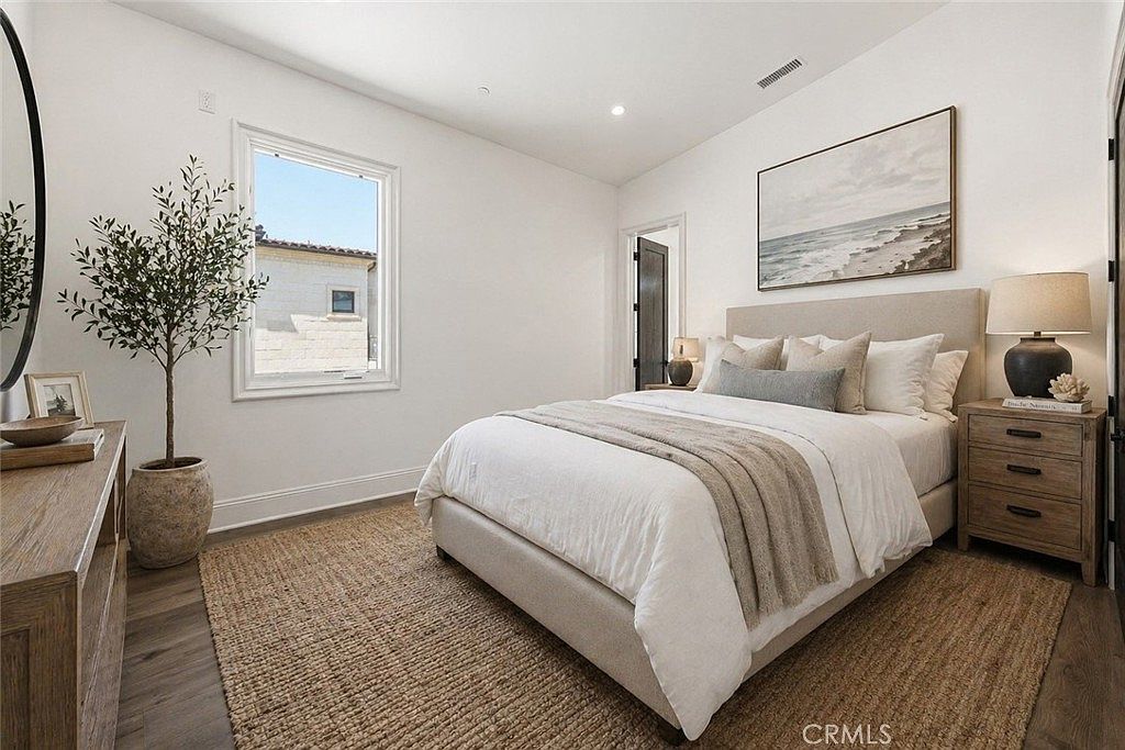 This bright and airy bedroom features a neutral color palette with a plush upholstered bed as the focal point. A large window allows for ample natural light, while a textured rug and a potted olive tree add warmth and organic texture to the space. The room is completed by a wooden nightstand with a table lamp and a coastal-themed landscape painting above the headboard.