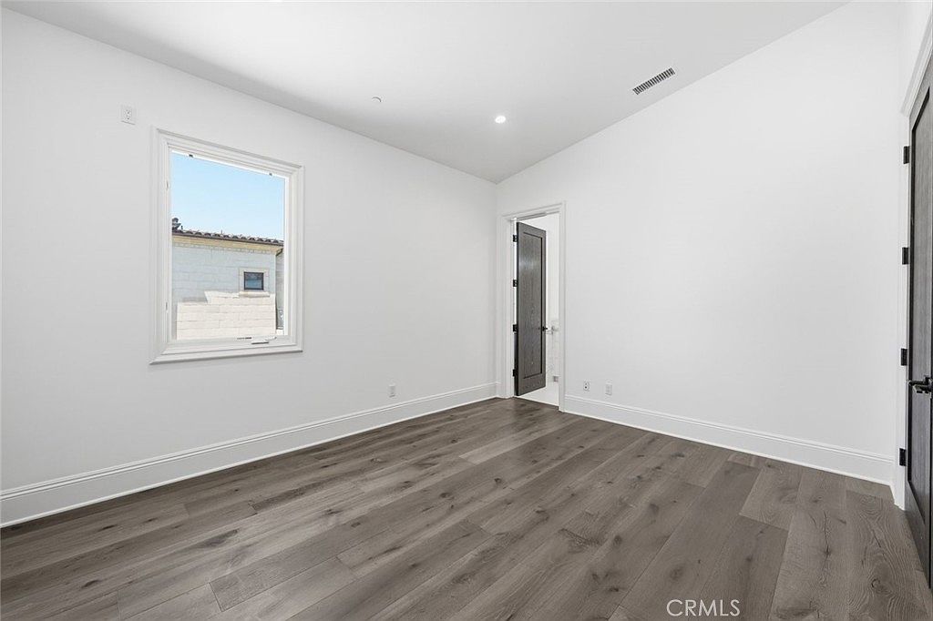 This bright, minimalist bedroom features crisp white walls and wide-plank wood-look flooring, creating a clean and airy atmosphere. A single window allows natural light to fill the space, while two dark-toned doors provide a sharp contrast to the neutral palette. The room is presented from a wide-angle perspective, emphasizing its simple, modern layout and potential for versatile use.
