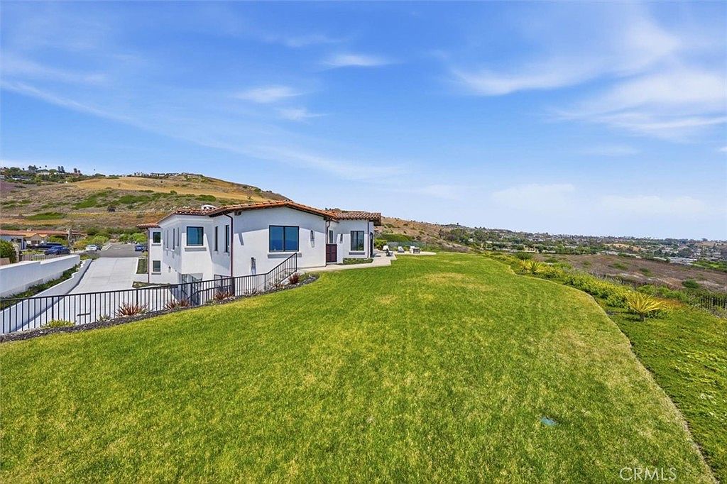 This image captures the rear exterior of a multi-level Mediterranean-style home, showcasing its white stucco facade and terracotta-tiled roof. A expansive, well-manicured lawn stretches out from the house, leading toward a scenic hillside landscape under a bright blue sky. The perspective is elevated, highlighting the property's generous outdoor space and its integration into the surrounding coastal terrain.