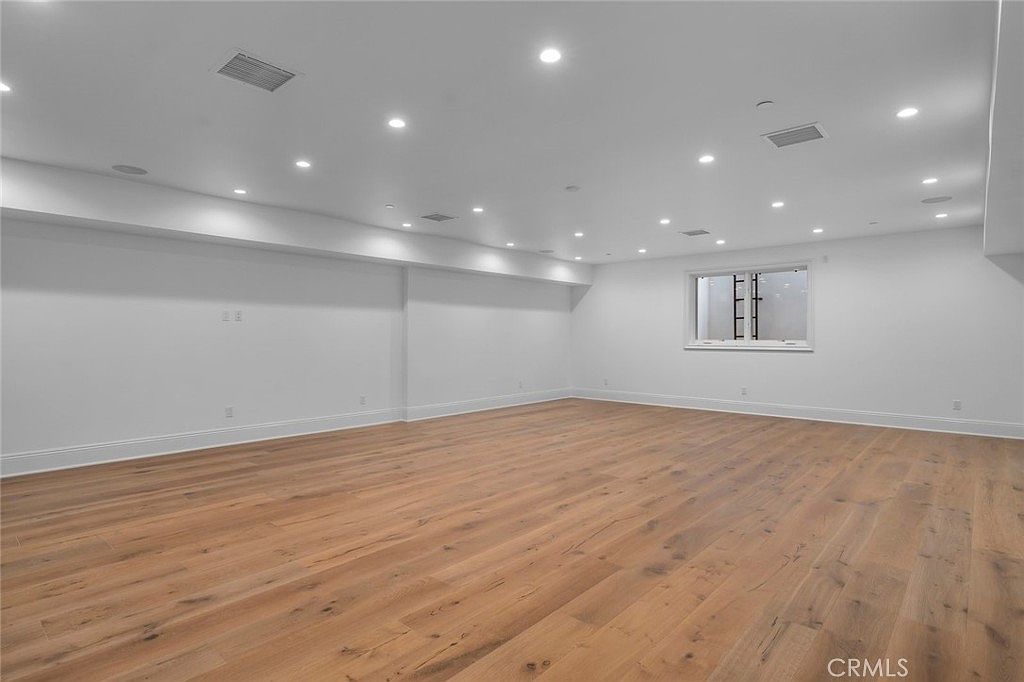This spacious, open-concept basement features light wood-toned flooring and crisp white walls, creating a bright and versatile living area. The room is illuminated by numerous recessed ceiling lights and includes a single window on the far wall. The clean, minimalist aesthetic provides a blank canvas suitable for a variety of uses, such as a recreation room or media space.