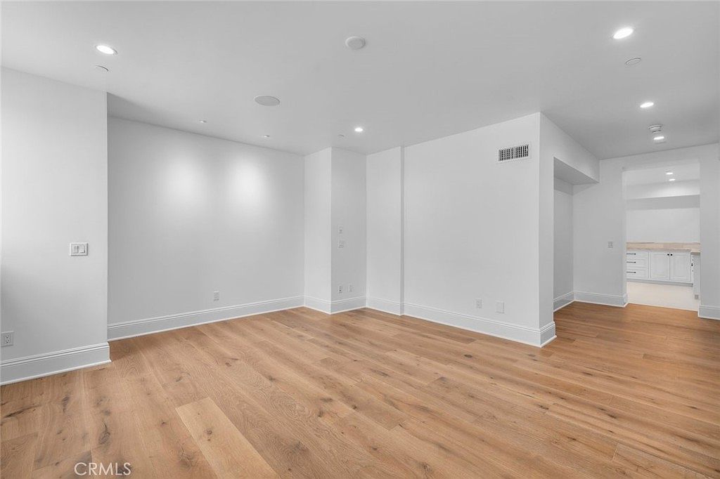This bright, open-concept living area features light wood flooring and crisp white walls, creating a clean and airy atmosphere. The space flows seamlessly into an adjacent kitchen area visible through a wide doorway, highlighting the home's modern, connected layout. Recessed lighting is integrated into the ceiling, and the room is framed by simple white baseboards, offering a versatile blank canvas for interior design.