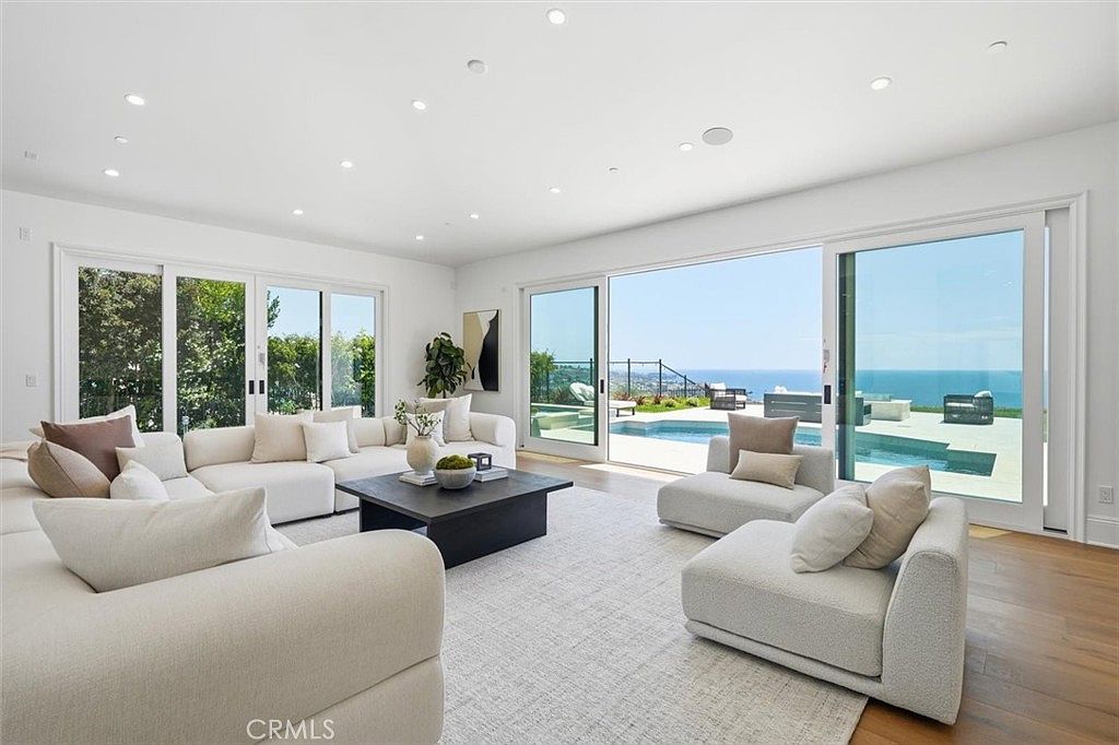 This bright and airy living room features a neutral color palette with plush, light-toned sectional seating arranged around a dark square coffee table. Large sliding glass doors open to a patio area, seamlessly blending the indoor space with the outdoor pool and ocean views. The room is illuminated by recessed lighting and natural light, creating a sophisticated and tranquil atmosphere.