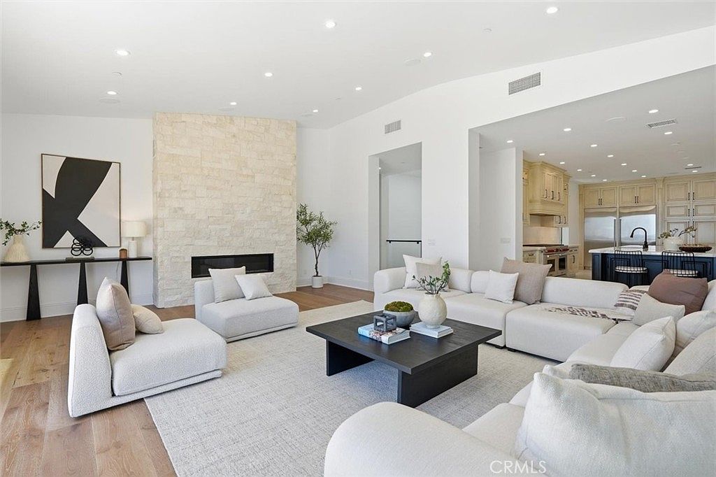 This spacious, open-concept living room features a neutral color palette and a large, light-toned sectional sofa arranged around a dark wood coffee table. A prominent stone-clad chimney breast serves as a focal point, housing a linear fireplace, while light wood flooring runs throughout the space. The room flows seamlessly into a bright kitchen area in the background, creating an airy and inviting atmosphere suitable for modern living.