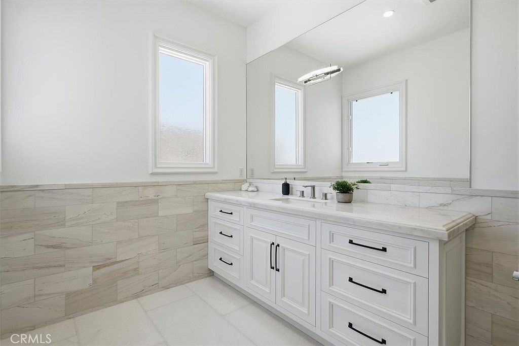 This bright and modern bathroom features a long white vanity with multiple drawers and a stone countertop. A large, wall-to-wall mirror reflects the space, which is accented by light-toned, horizontal-patterned wall tiles and neutral floor tiles. Two windows provide ample natural light, creating a clean and airy atmosphere.