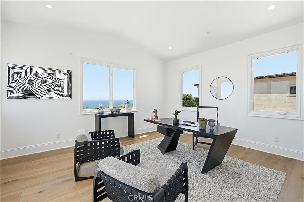 This bright and airy home office features light wood flooring and crisp white walls, creating a clean, modern aesthetic. The room is furnished with a sleek black desk and two contemporary woven chairs, while large windows provide ample natural light and a scenic view of the ocean. A large, abstract black-and-white art piece hangs on the wall, adding a bold focal point to the minimalist space.