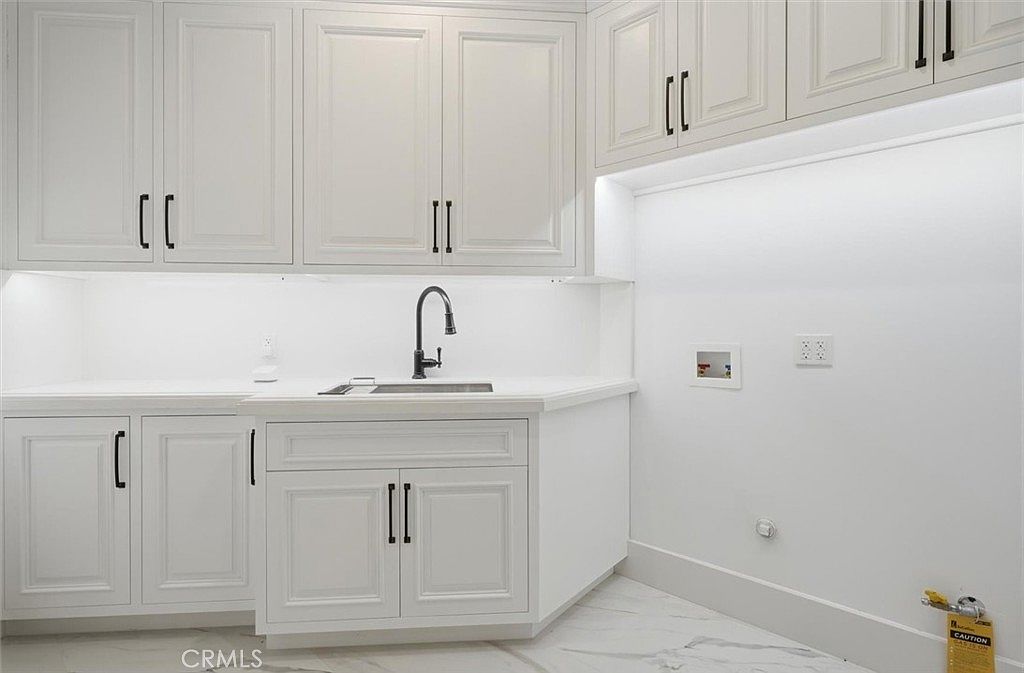 This clean and modern laundry room features crisp white cabinetry with ample storage space and a sleek, undermount sink with a dark faucet. The room is finished with light-toned, patterned flooring and bright under-cabinet lighting that illuminates the workspace. A dedicated area for a washer and dryer is visible, complete with necessary utility hookups and a gas line connection.