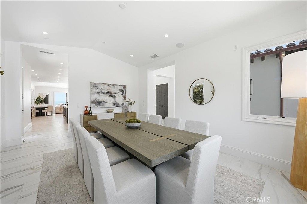 This bright and airy dining room features a long, light-toned wood table surrounded by ten upholstered chairs, set atop a neutral area rug. The space is characterized by white walls, light-colored tile flooring, and a minimalist aesthetic, with a large window providing natural light and a view of the outdoors. A piece of abstract wall art hangs above a sideboard, while a circular mirror adds a decorative touch to the adjacent wall.