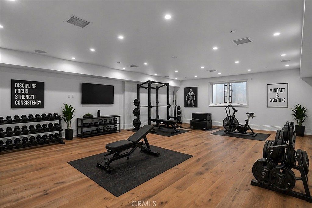This spacious, well-lit home gym features light wood-look flooring and a variety of professional-grade fitness equipment, including a squat rack, a weight bench, and a stationary bike. The walls are adorned with motivational signage and framed artwork, creating an energetic atmosphere. A wall-mounted television and ample recessed lighting complete this functional and modern workout space.