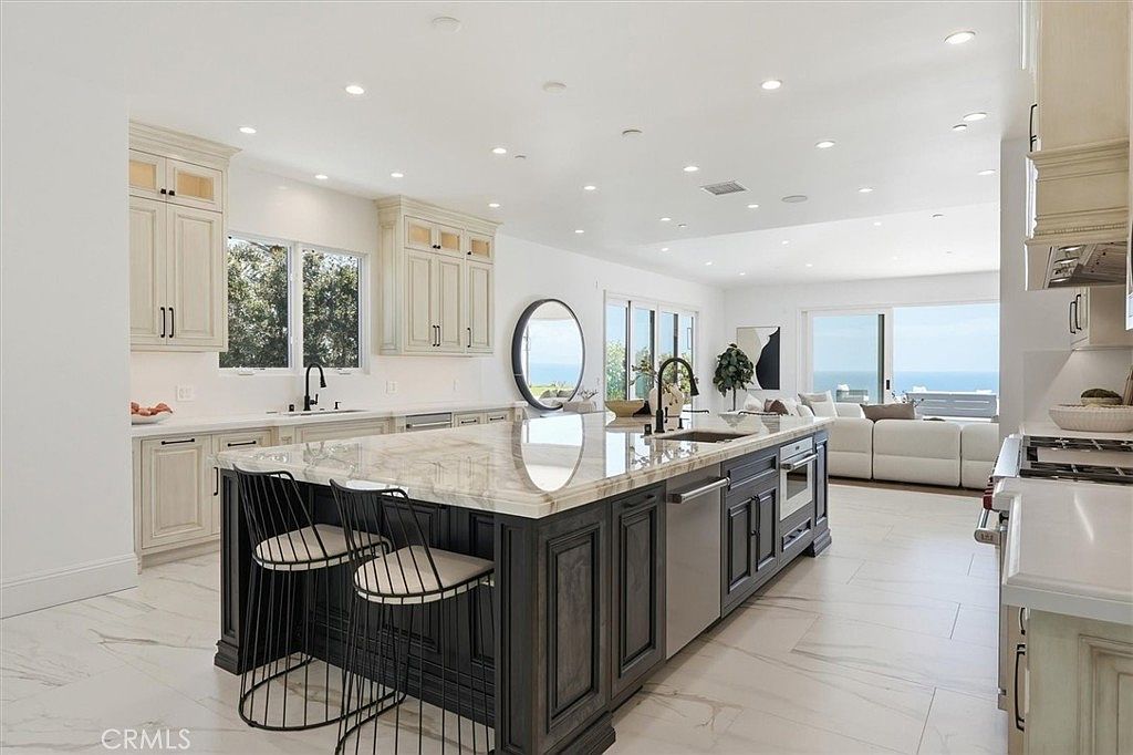This bright, open-concept kitchen features a large central island with a dark base and a light-toned stone countertop, complemented by light-colored cabinetry and recessed lighting. The space flows seamlessly into a living area with large sliding glass doors that reveal an expansive ocean view. The overall aesthetic is modern and airy, emphasizing a clean, high-end design with a neutral color palette.