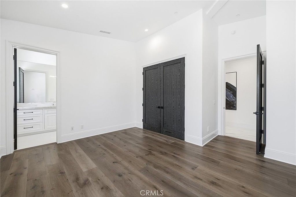 This bright, minimalist bedroom features wide-plank wood-look flooring and crisp white walls. The room includes a set of dark, double-panel doors, a doorway leading to an adjacent bathroom with a white vanity, and another open doorway leading to a hallway. The clean lines and neutral color palette create a versatile and modern living space.