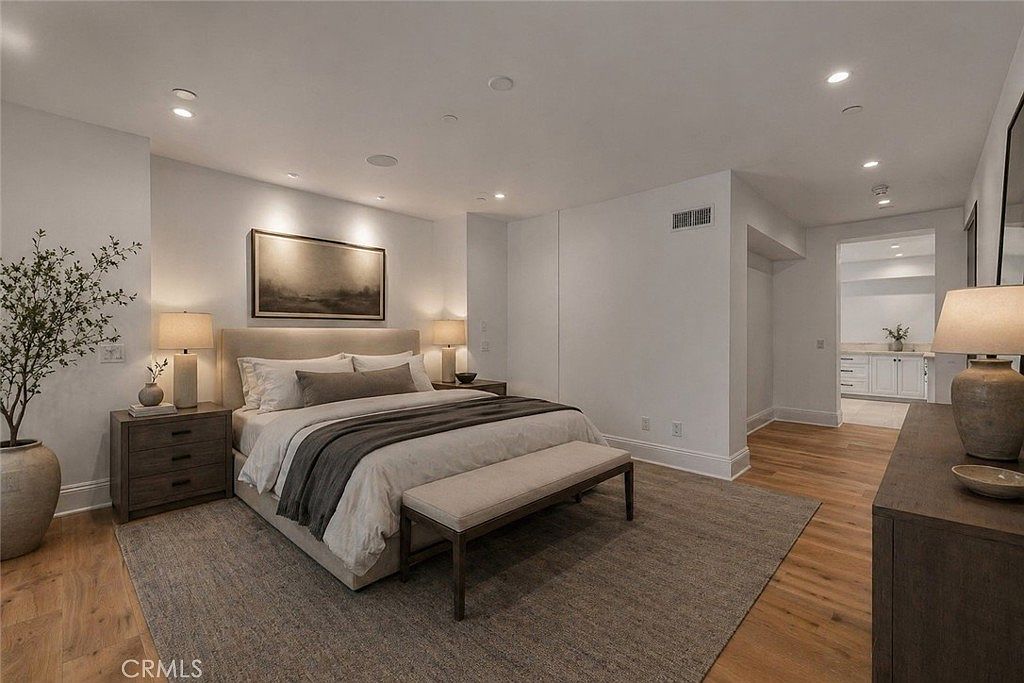 This serene primary bedroom features a neutral color palette with a large upholstered bed as the focal point, flanked by matching nightstands and table lamps. A long bench sits at the foot of the bed atop a textured area rug, while light wood flooring extends throughout the room. The space opens into an adjacent bathroom area, creating an airy and cohesive flow.