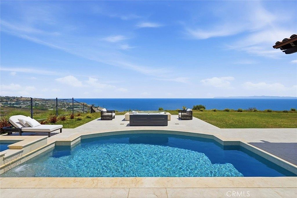This stunning outdoor space features a custom-shaped swimming pool with light-colored stone coping, set against a panoramic ocean view. A paved patio area includes a central fire pit with lounge seating, creating an ideal environment for relaxation and entertaining. The composition captures a serene, high-end coastal lifestyle with a clear horizon line and expansive sky.