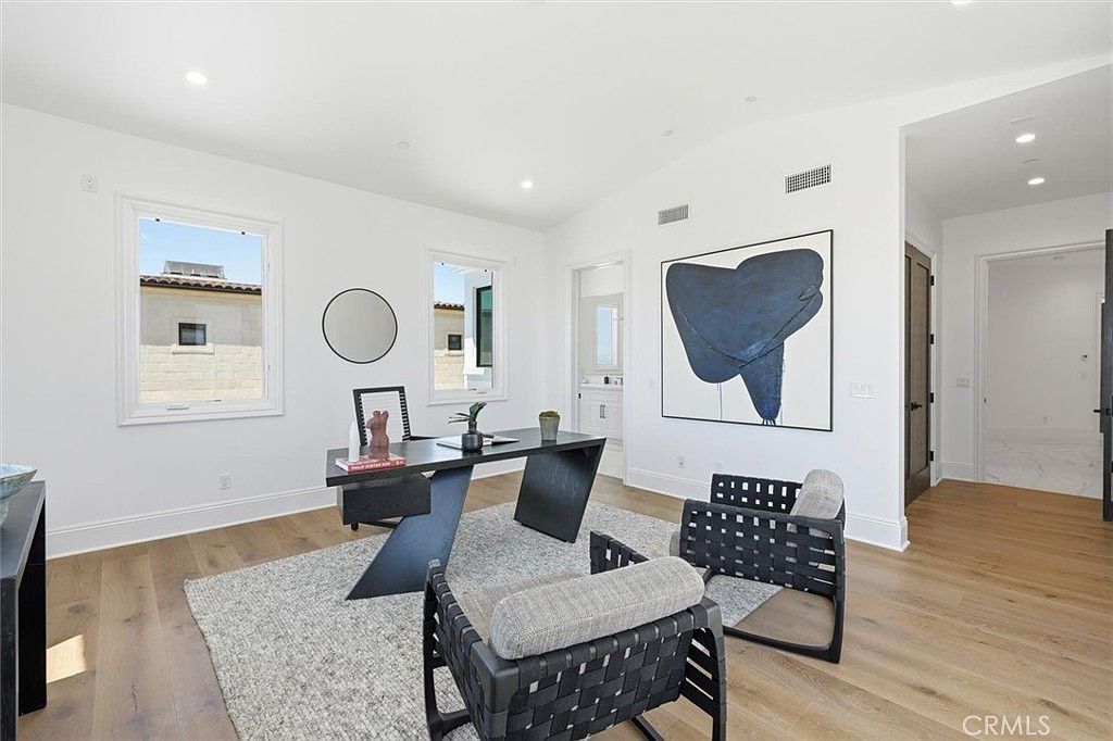 This bright and modern home office features light wood flooring, white walls, and a minimalist black desk positioned in the center of the room. Two contemporary black-framed chairs with woven seating face the desk, while a large abstract blue and black painting serves as a focal point on the wall. The room is illuminated by natural light from two windows and recessed lighting, creating a clean and professional atmosphere.