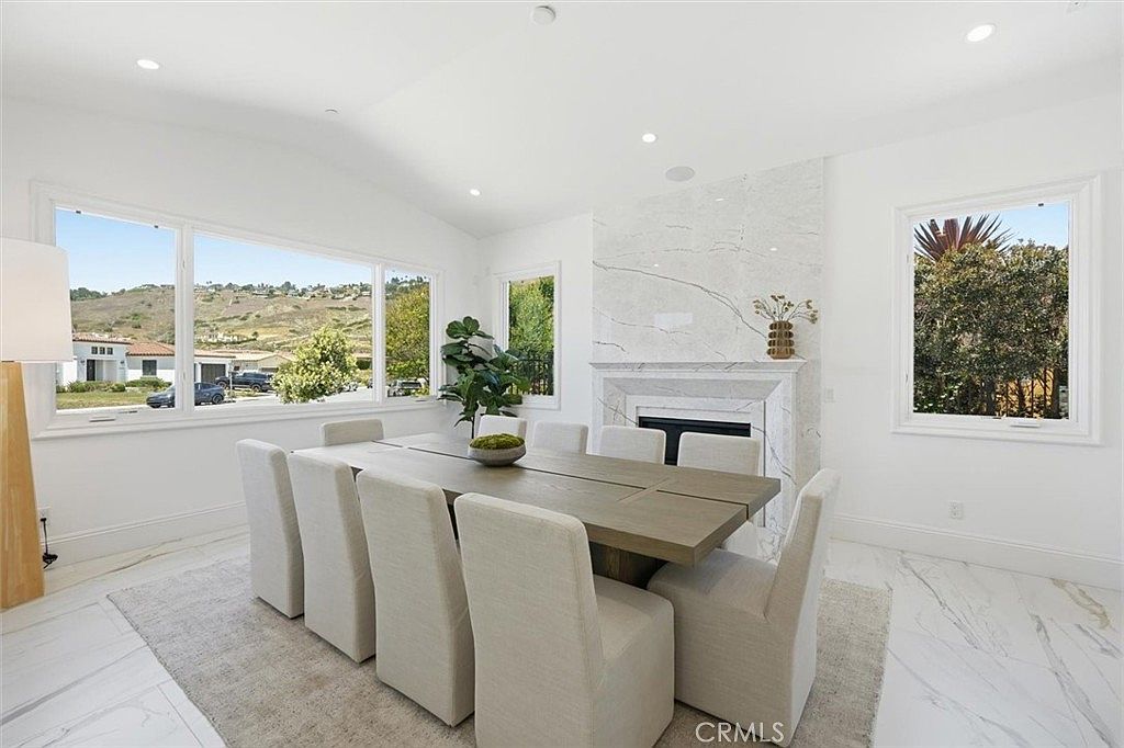 This bright and airy dining room features a large, light-toned wooden table surrounded by eight upholstered chairs. A prominent stone-clad chimney breast with a built-in fireplace serves as a focal point, while large windows provide ample natural light and views of the surrounding landscape. The space is finished with light-colored, patterned flooring and a neutral area rug, creating a sophisticated and welcoming atmosphere.