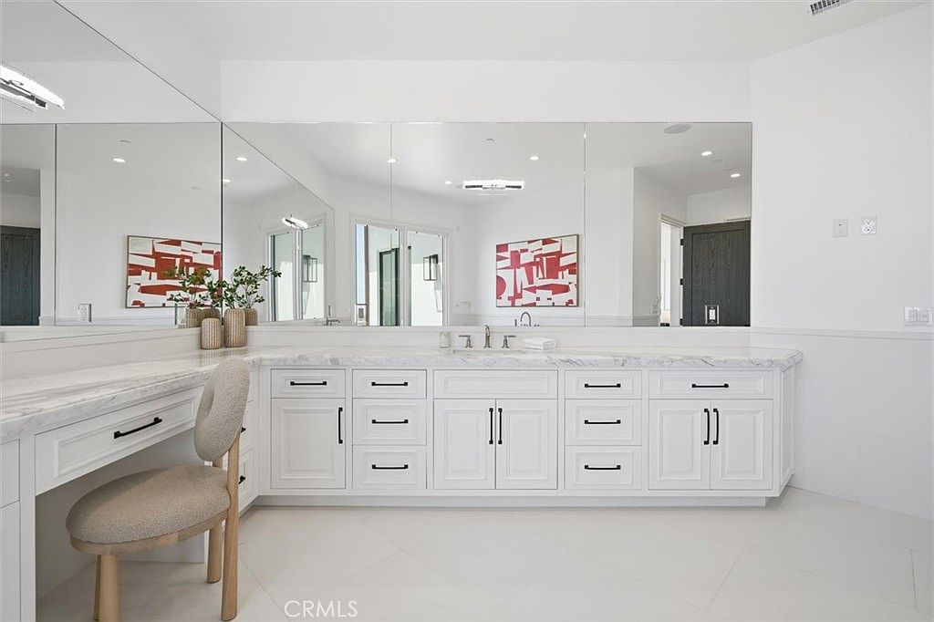 This bright and spacious primary bathroom features a long, white vanity with a light-toned stone countertop and a built-in makeup desk area. A large, expansive mirror spans the length of the wall, reflecting the clean, minimalist aesthetic and the neutral-toned chair. The room is finished with white cabinetry, modern hardware, and light-colored flooring, creating a polished and airy atmosphere.
