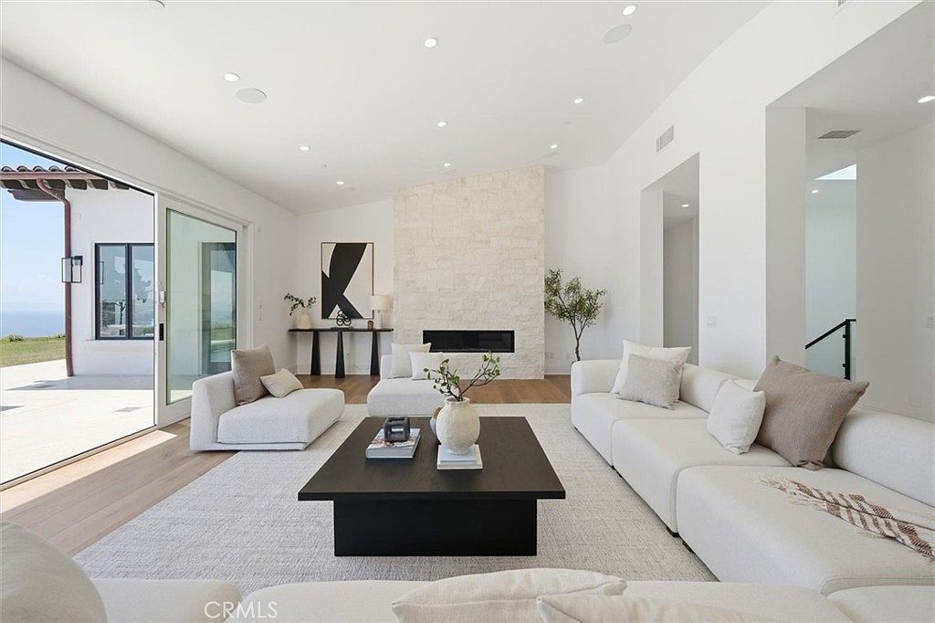 This bright and airy living room features a neutral color palette with plush white sectional seating arranged around a large, dark square coffee table. A prominent stone-clad chimney breast serves as the focal point, housing a sleek, linear fireplace. Large sliding glass doors open to an outdoor patio, providing an expansive view of the horizon and flooding the space with natural light.