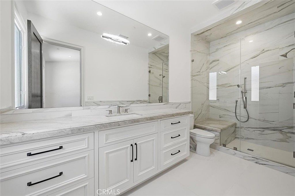 This bright, modern bathroom features a double vanity with white cabinetry, dark hardware, and a stone countertop. A large, frameless glass-enclosed shower with light-toned, veined stone wall tiles and a built-in bench sits adjacent to a white toilet. The space is illuminated by a sleek, contemporary light fixture above a wide mirror, creating a clean and luxurious atmosphere.