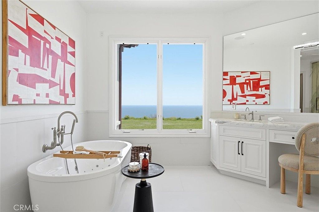 This bright, modern primary bathroom features a freestanding soaking tub positioned beneath a large window that offers a panoramic view of the ocean. To the right, a white vanity with a stone countertop and a large mirror provides a clean, minimalist aesthetic, complemented by a textured chair. The room is decorated with bold, abstract red and white artwork on the walls, creating a striking contrast against the neutral white interior.