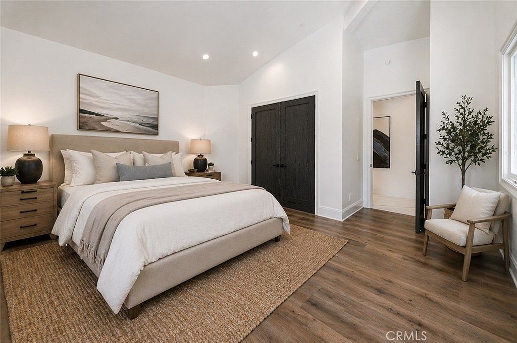 This bright and airy primary bedroom features a neutral color palette with a large bed centered against a wall adorned with a piece of landscape art. The room includes dark-toned double doors, warm wood-look flooring, and a cozy seating area with a light-colored armchair near a window. The space is illuminated by recessed lighting and two bedside lamps, creating a serene and inviting atmosphere.