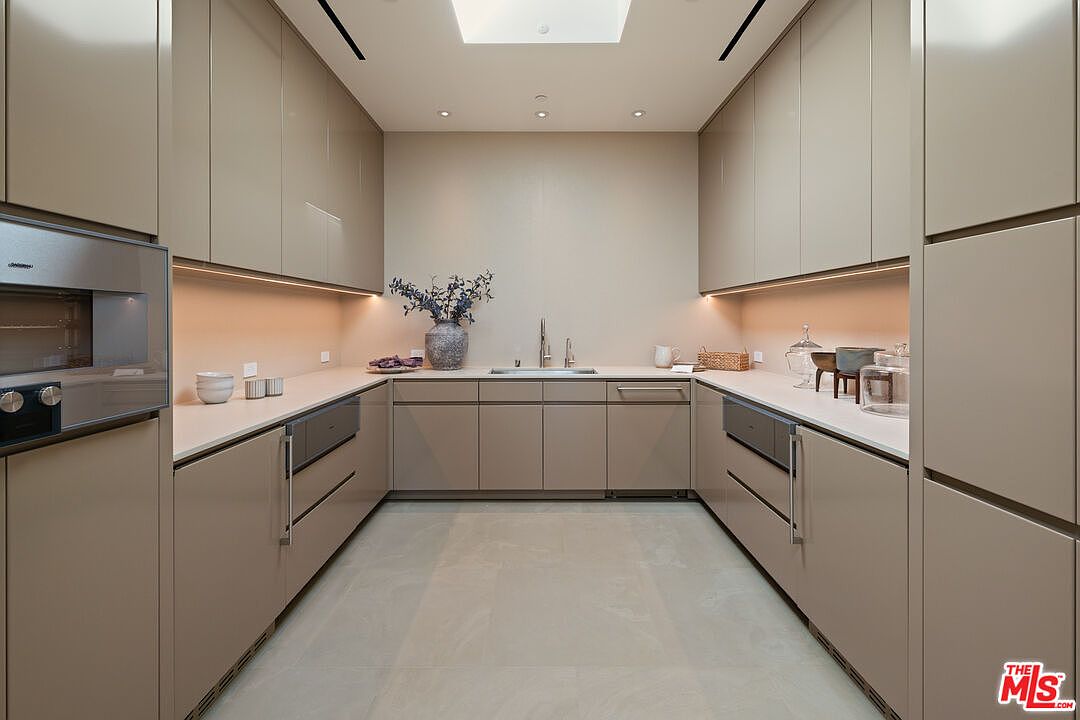This sleek, modern kitchen features a U-shaped layout with high-gloss, taupe-colored cabinetry and integrated appliances for a seamless, minimalist aesthetic. The space is illuminated by recessed lighting and a central skylight, creating a bright and airy atmosphere. A central sink area with a modern faucet serves as the focal point, complemented by clean, light-toned countertops and large-format floor tiles.