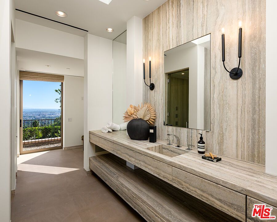 This luxurious primary bathroom features a stunning, custom-built stone vanity with a seamless integrated sink and a matching stone backsplash that extends to the ceiling. The space is accented by elegant, modern wall sconces and a large, frameless mirror, while a glass door leads to a balcony offering a breathtaking, expansive view of the city. The overall aesthetic is sophisticated and serene, utilizing natural textures and high-end materials to create a spa-like atmosphere.