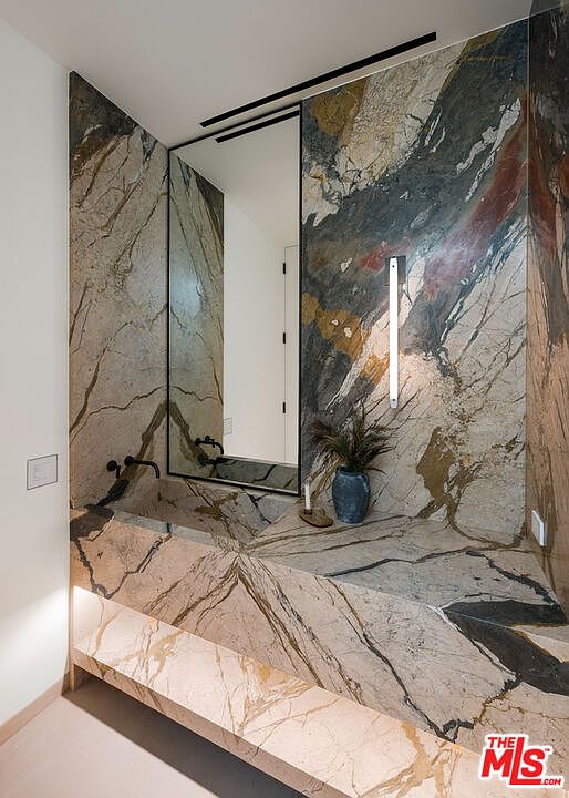 This sophisticated powder room features dramatic, book-matched marble slab walls and a matching integrated stone vanity. A sleek, vertical wall-mounted light fixture and a minimalist mirror create a modern, high-end aesthetic. The perspective is a straight-on shot that highlights the luxurious textures and clean lines of the space.