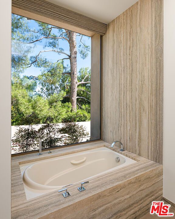 This luxurious bathroom features a deep soaking tub encased in elegant, vertically-veined travertine stone that extends up the adjacent wall. A large, floor-to-ceiling window provides a serene, unobstructed view of lush green trees and natural foliage, creating a spa-like atmosphere. The minimalist design and high-end stone finishes emphasize a sophisticated and tranquil retreat.