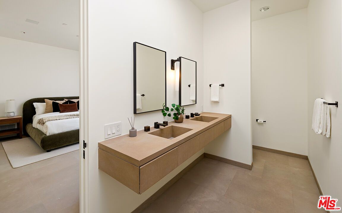 This modern primary bathroom features a sleek, floating double vanity with integrated concrete-style sinks and minimalist black-framed mirrors. The space is characterized by its clean lines, neutral color palette, and seamless transition into the adjacent bedroom. The perspective captures the vanity area from a slightly elevated angle, emphasizing the contemporary design and open-concept layout.