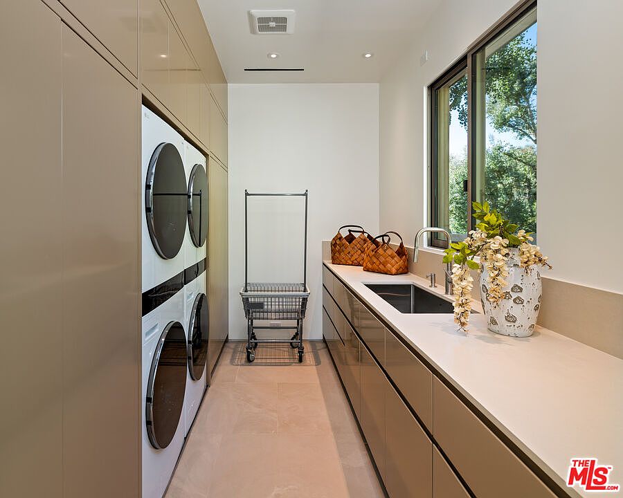 This sleek, modern laundry room features high-gloss taupe cabinetry and a long, clean countertop with an integrated sink. The space is equipped with a stacked washer and dryer unit, a rolling laundry cart, and a large window that brings in natural light, creating a bright and functional workspace. The minimalist design and neutral color palette give the room a sophisticated and organized aesthetic.