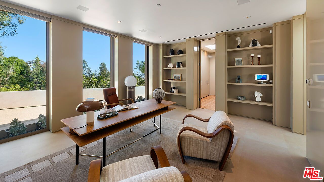 This sophisticated home office features a sleek, modern wooden desk paired with comfortable, mid-century inspired armchairs on a textured area rug. Large floor-to-ceiling windows flood the room with natural light, offering a serene view of the surrounding greenery, while built-in shelving provides elegant storage and display space. The neutral color palette and clean lines create a professional yet inviting atmosphere perfect for focused work.