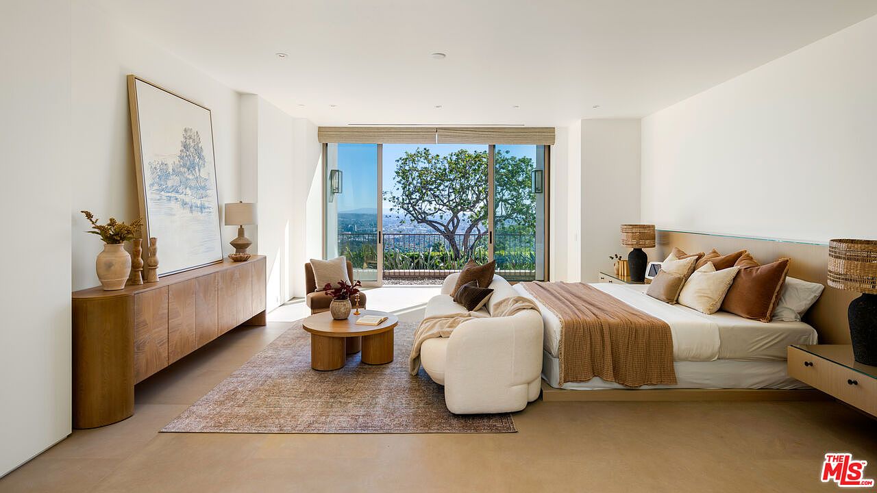 This spacious and serene primary bedroom features a contemporary design with a neutral color palette and warm wood accents. A plush, curved sofa sits at the foot of the bed, while large sliding glass doors open to a private balcony with stunning panoramic views. The room is elegantly furnished with a low-profile bed, a long wooden console, and textured decor, creating a sophisticated and tranquil atmosphere.