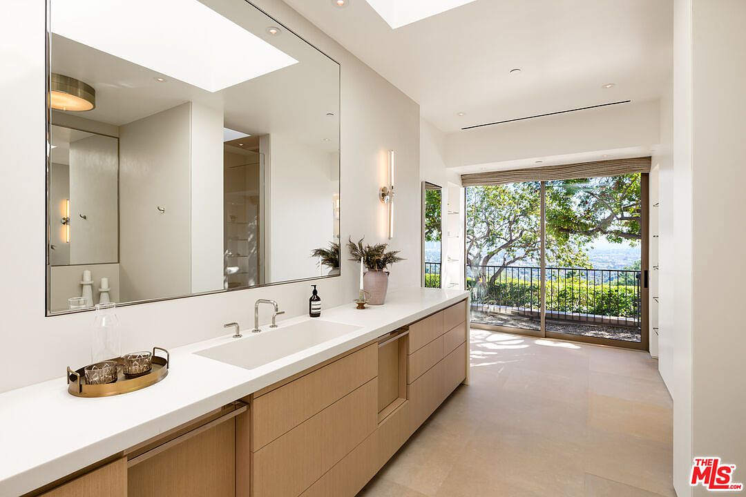 This modern primary bathroom features a sleek, light wood vanity with a long white countertop and a large, frameless wall-to-wall mirror. The space is brightened by natural light from a large skylight above and a sliding glass door that opens to a scenic outdoor view. The minimalist design, neutral color palette, and clean lines create a serene and luxurious atmosphere.