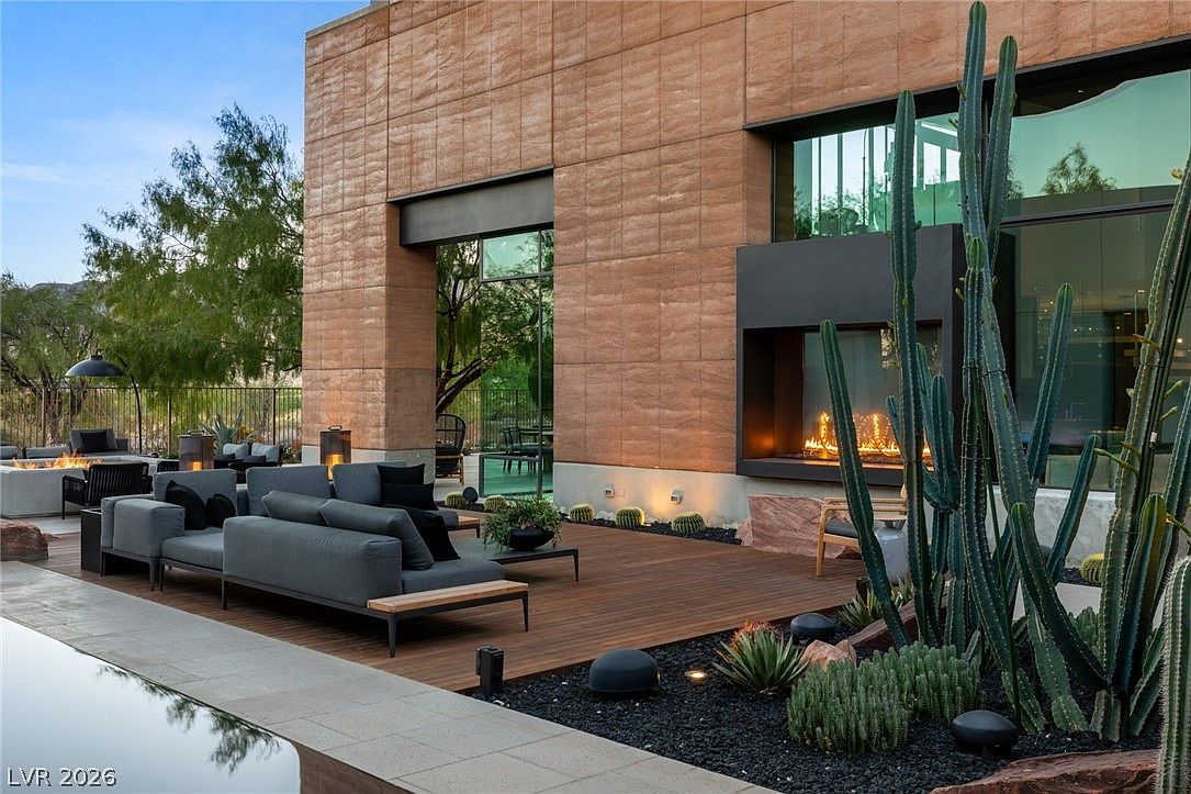 This sophisticated outdoor living space features a wooden deck furnished with a modern, low-profile sectional sofa and a central coffee table. A prominent outdoor fireplace with a tall, dark surround serves as a focal point, while a variety of desert cacti and dark gravel landscaping add texture to the scene. The perspective captures a seamless transition between the indoor living area and the pool-side patio, emphasizing a luxurious, contemporary desert aesthetic.
