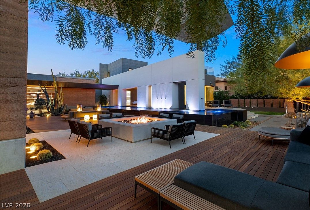 This luxurious outdoor living space features a multi-level deck with a central fire pit surrounded by modern lounge seating. A sleek, illuminated water feature serves as a dramatic backdrop, complemented by clean architectural lines and desert-inspired landscaping. The scene is captured at twilight, highlighting the warm glow of the fire and integrated landscape lighting against the deep blue evening sky.